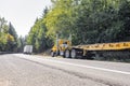 Convoy of two different big rig semi trucks with flat bed and dry van semi trailers driving on the winding narrow road with forest