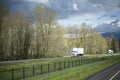 Convoy of the white big rigs semi trucks with semi trailers driving on the straight wide highway with bare trees on the side Royalty Free Stock Photo