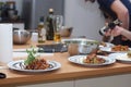 A cook in an apron sets the table for several people, laying out tomatoes in a caprese salad with pesto