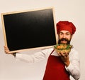 Cook with cheerful face in burgundy uniform hold baked dish and blackboard. Man with beard on white background. Chef