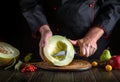 The cook cuts a large ripe melon with a knife on a wooden cutting board. Work environment with fruits on the kitchen table of a Royalty Free Stock Photo