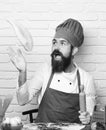 Cook with excited face in uniform sits by table with vegetables holding rolling pin. Man with beard throws pizza dough Royalty Free Stock Photo