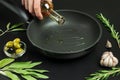 Cook hand pours olive oil from bottle into frying pan. Dark background with green and black olives, sprigs of rosemary Royalty Free Stock Photo