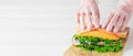 Cook`s hands lay a herring fillet sandwich with onions, cucumber and salad on paper. White wooden background with copy space for Royalty Free Stock Photo