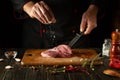 The cook sprinkles salt on the raw meat. Preparing beef steak before baking. Working environment in a restaurant or cafe kitchen Royalty Free Stock Photo