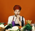 Cook works in kitchen near table with vegetables and tools. Chef with surprised face sits by food on red background Royalty Free Stock Photo