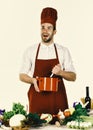 Cook works in kitchen near table with vegetables and tools. Man in cook hat and apron mixes in pot Royalty Free Stock Photo