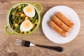 Vegetable mix, boiled egg in bowl, knife, salt shaker, pepper shaker, fried sausages in plate, fork on table. Top view Royalty Free Stock Photo