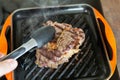 Cooking meat on cast iron grill pan indoors at home. Person turning ribeye steak on other side with tongs Royalty Free Stock Photo