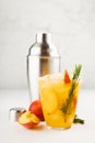 Cooking of refreshing juicy peach cocktail with ice, rosemary, sugar rim in misted glass, ingredients, silver shaker on white wood