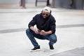 Cool African American man using smartphone, squatting on city street, wears black hoodie, jeans and sneakers, baseball Royalty Free Stock Photo