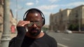 Cool afro american man with wireless headphones on head is walking to camera on street and putting on sunglasses