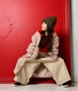 Cool asian kid girl in furcoat and wide pants is posing with her legs wide apart and hands on her knees looking aside Royalty Free Stock Photo