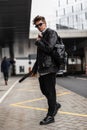 Cool attractive young man in sunglasses in an fashion oversized leather black jacket in jeans in boots with a stylish backpack Royalty Free Stock Photo