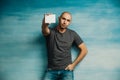 A cool bald man looks at the camera and holds out a white card, stands on a blue background, his second hand in his pocket Royalty Free Stock Photo