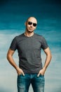 Cool bald man in a pilot`s sunglasses posing in a gray T-shirt holding his hands in his pockets against the background