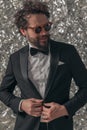 Cool bearded man in elegant tuxedo looking to side and fixing jacket