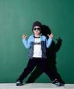 Cool boy in blue sunglasses, headwear, fleece jacket, pants and sneakers is posing like a gangster with two finger guns