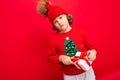 A cool boy with curls on a red wall background in a sweater with a Christmas tree holding a gift box in his hands Royalty Free Stock Photo