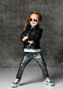 Cool brutal kid girl in leather jacket and shimmering jeans is standing with her legs wide apart and her hands crossed