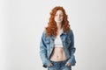 Cool brutal redhead girl in jean jacket chewing gum over white background.