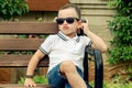 A cool child in sunglasses sits on a bench with his legs crossed.