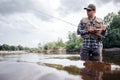 Cool fisherman stands in water and looks straight forward. He is serious. Guy holds fly rod and wooden box of artificial