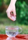 Cool water and ice cube in hand above the transparent glass cup in the table outoors in summer day.