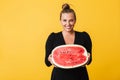 Cool girl in black dress happily winking in camera holding half of big watermelon in hands over yellow background
