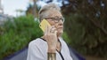 Cool, grey-haired senior woman caught in a serious smartphone conversation outdoors in sunny park, displaying a casual yet Royalty Free Stock Photo