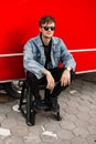 Cool handsome young man in a fashionable denim jacket in trendy black pants in vintage sunglasses resting sitting on a ladder near Royalty Free Stock Photo