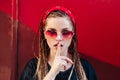 Cool hipster student woman wearing sunglasses and red bandana holds a finger on her mouth a secret on city street . Royalty Free Stock Photo