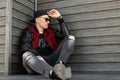 Cool hipster young man in trendy dark sunglasses in a vintage red and black checkered jacket with leather sleeves in jeans Royalty Free Stock Photo