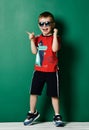 Cool kid boy in blue sunglasses sneakers, shorts and red t-shirt is laughing and pointing his finger at something Royalty Free Stock Photo