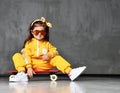 Cool kid girl in yellow hooded jumpsuit costume, sunglasses and yellow bow sits on skateboard in cool pose like just after trick