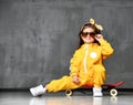 Cool kid girl in yellow hooded jumpsuit costume, sunglasses and yellow bow sits in cool pose on red skateboard