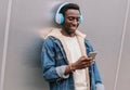 Cool modern smiling young african man in wireless headphones listening to music holding phone on city street over gray metal Royalty Free Stock Photo