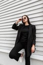 Cool modern young woman in fashionable black casual wear with handbag straightens sunglasses near vintage wooden building on Royalty Free Stock Photo