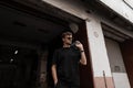 Cool nice young man in stylish sunglasses in a vintage blue denim jacket in a black T-shirt poses near a building on the street.