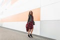 Cool positive young girl in red youth checkered dress in vintage leather jacket in trendy sunglasses in sneakers with bag travel Royalty Free Stock Photo