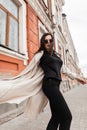 Cool pretty young woman hipster in vintage sunglasses in black stylish casual wear in spring fashion trench coat whirls outdoors Royalty Free Stock Photo