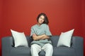 Cool skeptical european young brunette girl sitting with crossed arms on modern couch in studio, watching television Royalty Free Stock Photo