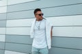 Cool young American man hipster in sunglasses in blue trendy jeans in a fashionable white t-shirt is posing in the city near a Royalty Free Stock Photo