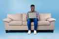 Cool young black guy sitting on sofa with laptop, working or studying online over blue studio background, copy space Royalty Free Stock Photo