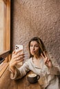 Cool young caucasian girl grimaces making selfie on smartphone sitting in cafe. Royalty Free Stock Photo