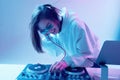 Cool young girl DJ mixes music on a mixing console and laptop, in stylish clothes, glasses on a neon background.