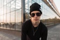Cool young handsome hipster man in stylish knitted black hat in trendy sunglasses in a t-shirt posing Royalty Free Stock Photo