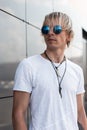 Cool young hipster man with blond hair in blue round sunglasses in a fashionable white t-shirt with amulets on a neck is standing Royalty Free Stock Photo