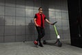 Cool young hipster man in fashionable denim red-black clothes in stylish sunglasses stands with a modern electric scooter Royalty Free Stock Photo