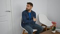 Cool young hispanic man, arms crossed in a relaxed gesture, sitting in a waiting room chair with a serious expression, looking Royalty Free Stock Photo